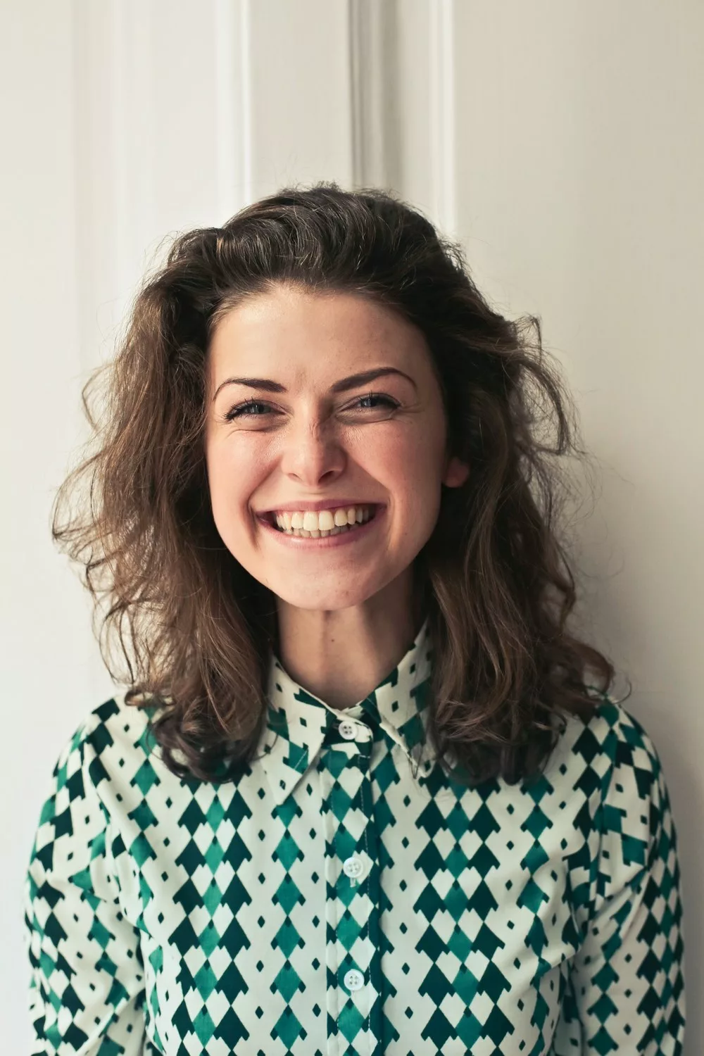 L'image montre une personne aux cheveux bruns bouclés, souriante et regardant directement l'appareil photo. Elle porte une chemise à motifs verts et blancs. L'arrière-plan semble être un mur uni de couleur claire.